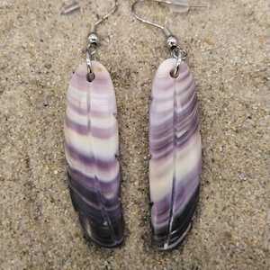 Puede incluir: Un par de pendientes colgantes con colgantes alargados en forma de concha. Los colgantes muestran un degradado de colores, incluyendo blanco, lavanda, morado y negro. Los pendientes tienen ganchos plateados y se exhiben sobre una superficie arenosa.