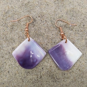 Puede incluir: Un par de pendientes con ganchos de color cobre y colgantes en forma de diamante. Los colgantes son blancos y morados, con un efecto veteado. Los pendientes se exhiben sobre una superficie arenosa.