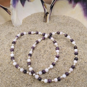 May include: Two bracelets made of white and purple shell beads with silver accents. The bracelets are lying on a bed of sand.