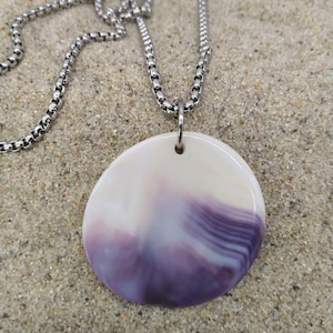 May include: A round pendant necklace with a silver chain. The pendant is white with a gradient of purple and lavender hues. The necklace is displayed on a sandy surface.
