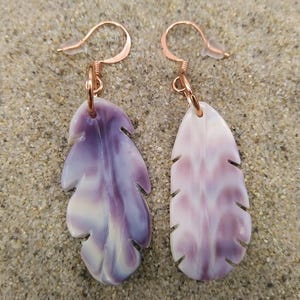 Puede incluir: Un par de pendientes con forma de pluma. Un pendiente es de color púrpura intenso con remolinos blancos, el otro es de color rosa claro con remolinos blancos. Ambos tienen ganchos para las orejas de color cobre y están sobre arena.