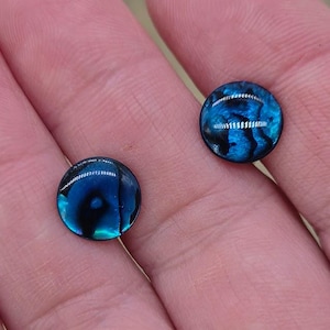 May include: Two round, iridescent blue cabochons with black accents. The cabochons have a glossy finish. These could be used for jewellery making or other craft projects. The cabochons are set against a neutral background.