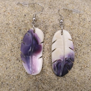 May include: A pair of feather-shaped earrings. Each earring has a gradient of white to purple hues. The earrings have silver hooks and are displayed on a sandy surface, suggesting a beach or coastal theme.