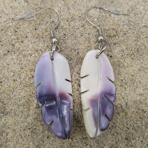 Puede incluir: Pendientes con forma de pluma en blanco y morado, con ganchos plateados. Los pendientes están sobre arena. El diseño de la pluma presenta líneas talladas. Miden aproximadamente 5 cm de largo.