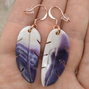 Puede incluir: Un par de pendientes en forma de pluma. Cada pendiente presenta un diseño veteado blanco y morado. Los pendientes tienen ganchos de color oro rosa y cierres de plástico transparente. Los pendientes se sostienen en una mano.