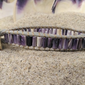 Puede incluir: Una pulsera artesanal con cuentas rectangulares moradas, negras y blancas. Las cuentas están ensartadas en un cordón de cuero gris y aseguradas con un cierre de madera. La pulsera se exhibe sobre un lecho de arena.