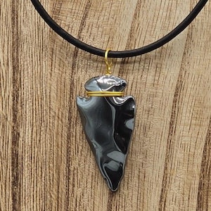 May include: A necklace featuring a dark gray, polished arrowhead-shaped pendant suspended from a black cord. The pendant is secured with thin gold-colored wire wrapped around the top notch, connecting to a small gold-colored metal jump ring.