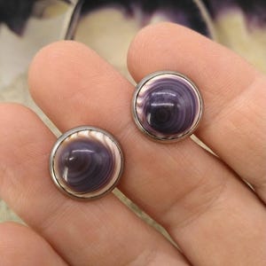 May include: A pair of round, silver-toned earrings with a polished, swirling purple and white design. The earrings have a domed, shell-like appearance, with a metallic border. The background features a blurred seashell.