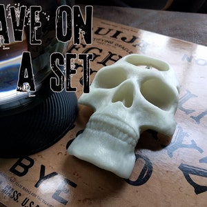 Könnte beinhalten: Ein weißer Plastik-Schädel mit einem Loch oben, der auf einem braunen Ouija-Brett mit den Buchstaben "BYE" liegt. Der Text "SAVE ON A SET" ist in der oberen linken Ecke des Bildes sichtbar.