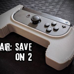 May include: A white and gray gaming controller grip with a black controller. The grip has the text "PAIR: SAVE ON 2". The controller has black buttons and a joystick. The grip is designed to enhance the gaming experience.
