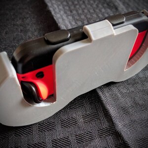 May include: A gray plastic controller grip encasing a black and red game controller. The grip has a curved design with a cutout revealing the controller's buttons. A black lanyard is attached.