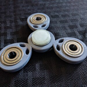 May include: A gray plastic fidget spinner with three arms and three ball bearings. The center of the spinner has a white plastic disc with a grid pattern.