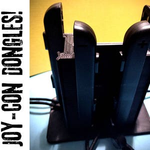 May include: Black Joy-Con controller holder with the text "HOLDS 8 JOY-CON DONGLES!" The holder has slots for multiple controllers and is designed to keep them organized. The holder is on a blue surface.