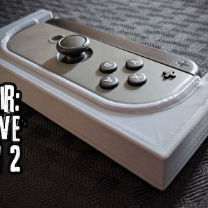 May include: A white plastic holder for a gray Nintendo Switch Joy-Con controller. The text "PAIR: SAVE ON 2" is visible in the image.