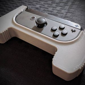 May include: A white 3D printed stand for a Nintendo Switch Joy-Con controller. The stand has a textured surface and a raised lip to hold the controller in place.