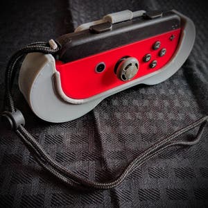 May include: A red and black game controller with gray accents and a black wrist strap. The controller has a gray protective case and features several black buttons and a gray joystick. The wrist strap is black and attached to the controller.