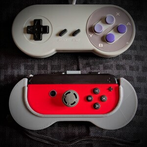 May include: Two video game controllers are displayed. The top controller is gray with black and purple buttons, labeled "SELECT" and "START." The bottom controller is red and gray with black buttons. Both controllers are on a textured black surface.