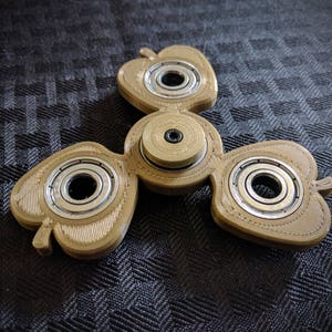 Puede incluir: Un spinner de dedos de tres puntas de color marrón con un diseño en forma de corazón. Cada punta tiene un rodamiento plateado en el centro.