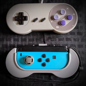 Può includere: Due controller per videogiochi. Il controller superiore è grigio con pulsanti viola e un pad direzionale. Il controller inferiore è blu e nero con accenti e pulsanti grigi. Entrambi i controller sono su una superficie scura a motivi.