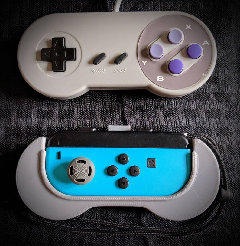May include: Two video game controllers. The top controller is gray with purple buttons and a directional pad. The bottom controller is blue and black with gray accents and buttons. Both controllers are on a dark patterned surface.