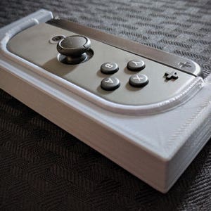 Op de afbeelding: Een 3D-geprinte witte kunststof houder voor een Nintendo Switch Joy-Con-controller. De houder is ontworpen om de controller veilig op zijn plaats te houden en heeft een uitsparing voor de knoppen van de controller.