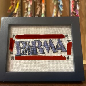 Op de afbeelding: Een ingelijst kruissteek met het woord "PARMA" in blauwe letters op een witte achtergrond. Het woord wordt omringd door twee rode strepen.