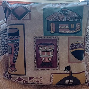 Könnte beinhalten: Quadratisches Zierkissen mit afrikanischem Stammesmuster. Das Kissen zeigt Bilder von Trommeln, Hütten und geometrischen Formen in Braun-, Beige- und Blautönen. Das Kissen liegt auf einem Korbstuhl.