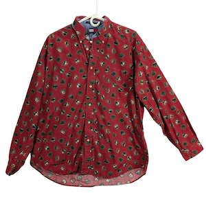 Op de afbeelding: Een rood overhemd met knopen en een herhalend schildpatroon in groen, zwart en bruin. Het shirt heeft lange mouwen en een button-down kraag. Het shirt hangt aan een witte hanger.