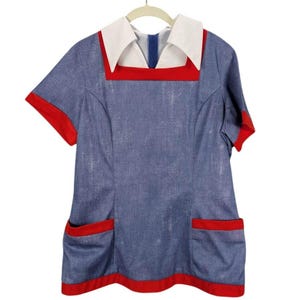 May include: A blue dress with red trim and white collar. The dress has short sleeves and two front pockets. The collar is square and the dress is hanging on a white hanger.
