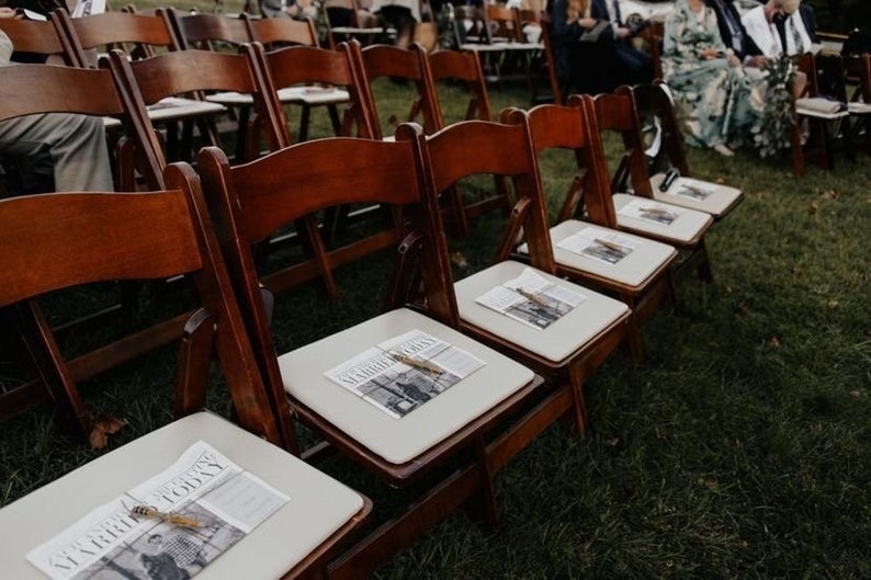 Puede incluir: Filas de sillas plegables de madera con cojines color crema y peri&oacute;dicos, dispuestas sobre hierba. Los peri&oacute;dicos est&aacute;n atados con hilo. Las sillas est&aacute;n preparadas para un evento al aire libre, como una boda.