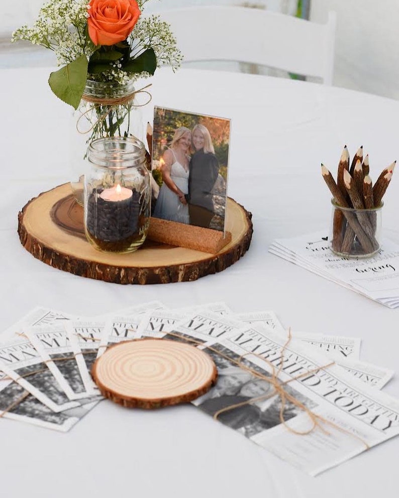 Puede incluir: Centro de mesa de boda con base de rodaja de madera, vela en tarro y foto enmarcada. Un peque&ntilde;o ramo de rosas naranjas y flores blancas est&aacute; en un tarro. Los programas de boda estilo peri&oacute;dico y los l&aacute;pices completan la decoraci&oacute;n.