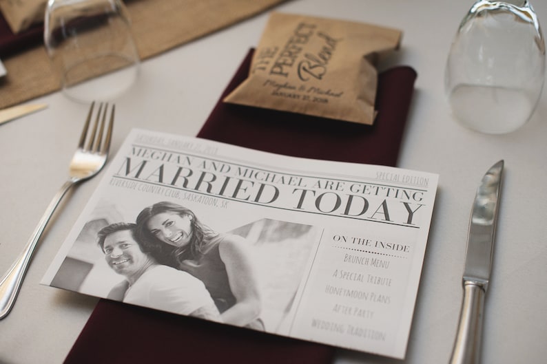 Puede incluir: Una mesa de recepci&oacute;n de bodas con un anuncio estilo peri&oacute;dico que dice "MEGHAN AND MICHAEL ARE GETTING MARRIED TODAY". La mesa tambi&eacute;n incluye un peque&ntilde;o paquete de caf&eacute;, cubiertos y copas de vino.