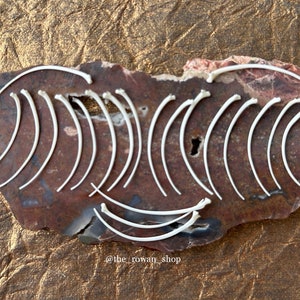 May include: A collection of white bone ribs arranged in a curved pattern on a brown and pink stone. The stone has a natural, rough texture. The text "@the_rowan_shop" is written in black at the bottom of the stone.