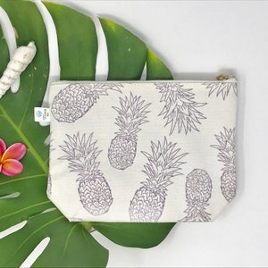 Puede incluir: Una bolsa de lona blanca con cremallera con un patrón repetido de piñas grises. La bolsa tiene una cremallera dorada.