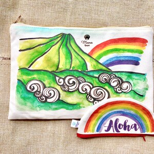 May include: A white canvas zippered pouch with a watercolour illustration of a green mountain with a rainbow arching over it. The pouch has a smaller matching rainbow pouch with the word "Aloha" written in purple on it.