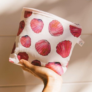 Op de afbeelding: Een witte make-up tas met een roze rits en een patroon van roze zeeschelpen. De tas wordt in een hand gehouden en lijkt gemaakt van canvas of een vergelijkbare stof. De zeeschelpen hebben een aquareleffect.