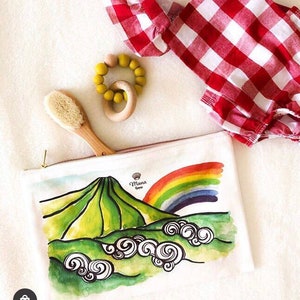 May include: White zippered pouch with a watercolour illustration of a green mountain, rainbow, and clouds. The text "Mana Hawaii" is printed on the pouch.