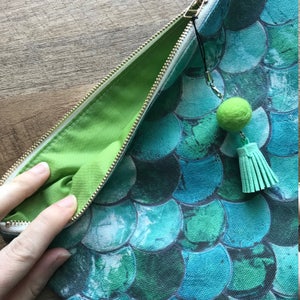 May include: A green zippered pouch with a green pom-pom tassel. The pouch is made of canvas and has a mermaid scale pattern in shades of blue and green.