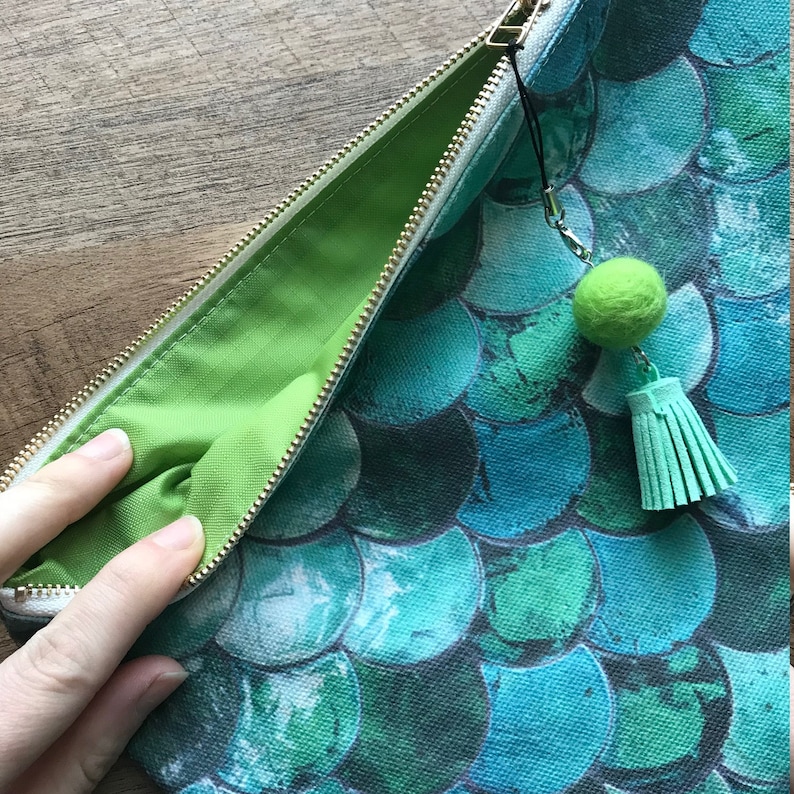 May include: A green zippered pouch with a green pom-pom tassel. The pouch is made of canvas and has a mermaid scale pattern in shades of blue and green.