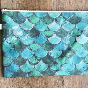 May include: A teal blue zippered pouch with a mermaid scale pattern. The pouch has a white zipper and a small green tassel.