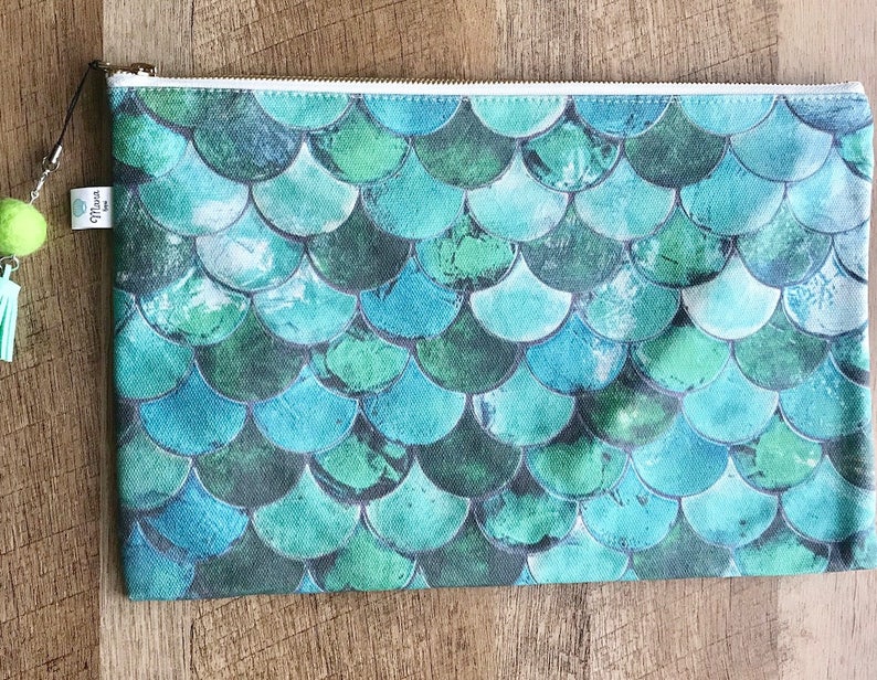 May include: A teal blue zippered pouch with a mermaid scale pattern. The pouch has a white zipper and a small green tassel.