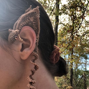 May include: A brown crocheted ear cuff and earring set. The ear cuff is designed to cover the ear, while the earring dangles in a spiral shape. A gold-colored hoop earring is also visible. The background shows a blurred view of trees.