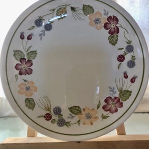 May include: A white ceramic dinner plate with a floral design. The plate has a green, yellow, and purple floral pattern with blackberries around the edge.