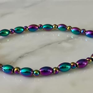 May include: A beaded bracelet with iridescent oval beads in shades of purple, teal, and gold. The bracelet is strung on an elastic band, and the beads have a metallic sheen. This jewelry item is suitable for everyday wear.