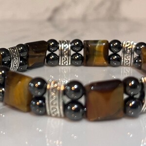 May include: A brown and gold tiger's eye stone bracelet with black hematite beads and silver spacers. The bracelet has a decorative design on the spacers.