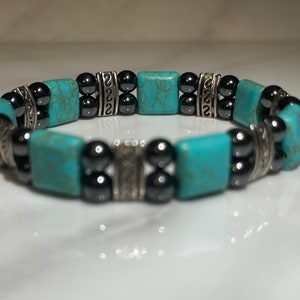 May include: A turquoise and black beaded bracelet with silver accents. The bracelet is made with square turquoise stones and round black beads.