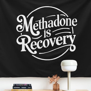May include: Black wall tapestry with the text "Methadone is Recovery" in a white, stylized font. The tapestry is hanging on a white wall above a wooden dresser. A lamp and decorative items are on the dresser.