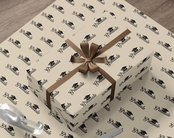 May include: A brown gift box wrapped in beige wrapping paper with a black and white pattern of graduation caps and the word "Graduate". A brown ribbon is tied around the box with a bow on top. A pair of silver scissors is lying on the wrapping paper.