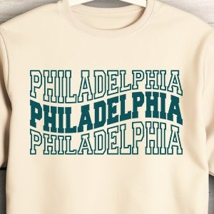 Może przedstawiać: Kremowa bluza z napisem "PHILADELPHIA" powtórzonym trzy razy w kolorze turkusowym. Tekst jest w retro, pogrubionej czcionce.