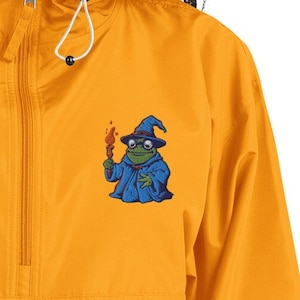 May include: Mustard yellow anorak jacket with a half-zip closure and a hood. The jacket features a small embroidered patch of a frog dressed as a wizard, holding a torch. The frog is wearing a blue robe and a wizard hat.